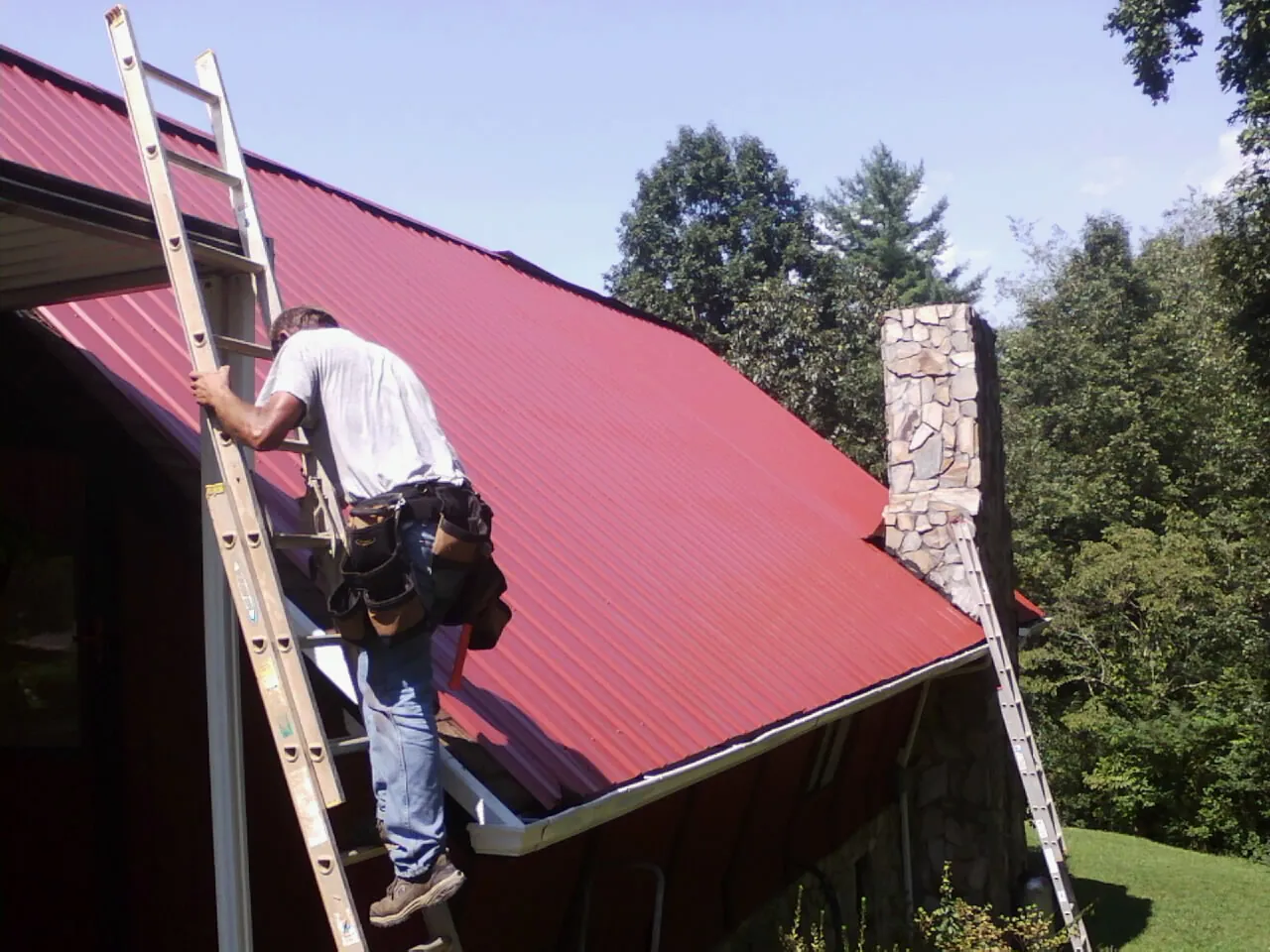 Roofer on a ladder beside a red metal roof and stone chimney — Soffit & Fascia Repair by Roofing Brothers in Easley